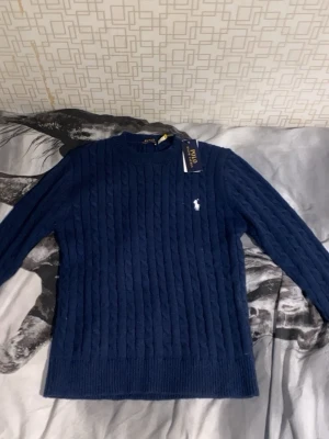 Marinblå kabelstickad tröja Polo Ralph Lauren - Kabelstickad marinblå tröja från Polo Ralph Lauren med rund halsringning och diskret logga broderad på bröstet. Tröjan har långa ärmar och ribbade muddar vid ärmslut och nederkant. Perfekt för lager på lager under kyliga dagar.