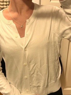 Vit kofta med knappar - Säljer en klassisk vit kofta med rund hals och knäppning framtill. Koftan har långa ärmar, ribbade muddar och är i ett mjukt, tunt material som känns skönt mot huden. Perfekt att slänga över en topp när det är lite kyligt ute. Den är lite nopprig men syns inte 