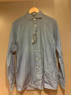 Blå skjorta från Eton i bomull - Snygg blå skjorta från Eton i storlek M med klassisk krage och ljusa knappar. Skjortan är tillverkad i mjuk bomull och har en stilren, tidlös look som passar perfekt till jeans eller chinos. Märkeslapp kvar på plagget.