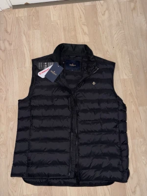 Svart dunväst från Morris - Säljer en svart quiltad dunväst från Morris med diskret logga på bröstet och hög krage. Västen har dragkedja framtill och är tillverkad i syntetmaterial med Primaloft-fyllning. Perfekt för lager-på-lager under höst och vinter. Helt ny med lapparna kvar! Nypris 2299 kronor.