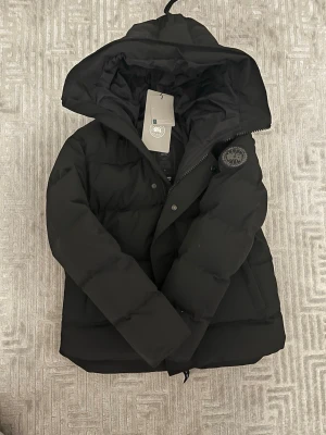 Svart Canada goose 165-175cm - Svart pufferjacka från Canada Goose i storlek L. Jackan har huva, dragkedja och knappar framtill samt ett broderat märke på ärmen. Fylld med dun och har en clean, oversized look som är perfekt för kalla dagar. Snygg och funktionell design med rejäla fickor.