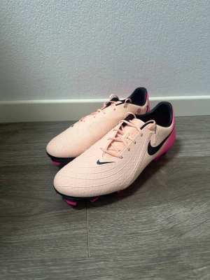Nike fotbollsskor i ljusrosa och neonrosa - Säljer ett par riktigt snygga Nike fotbollsskor i ljusrosa med neonrosa sula och svarta detaljer. Skorna har snörning, rund tå och är tillverkade i syntetmaterial för bästa grepp på planen. Perfekta för dig som vill sticka ut på matchen!