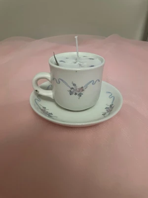 Doftljus av vaniljdoft i en vit tekopp med blommor och blåa streck nr 1  - Det här ljuset är gjort av återanvändbara material. Tekoppen är köpt secondhand och stearinet är från kyrkor som tänkte slänga det. Det här ljuset har en svag doft utav vanilj, så känsliga även kan använda det. Det här ljuset har även torkade blommor på som dekoration. Det finns fler av denna design på hemsidan om man vill köpa sets. 