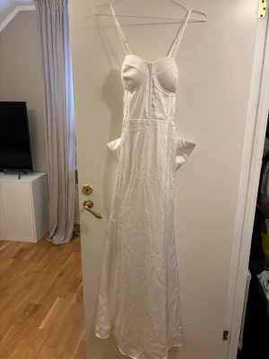 Vit långklänning med rosett från Boohoo - Supersnygg vit långklänning från Boohoo i glansigt satinliknande material. Klänningen har smala axelband, markerad byst och en stor rosett bak som ger en extra wow-effekt. Perfekt för dig som vill sticka ut och känna dig extra glam.