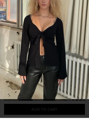 Svart långärmad topp med knyt - Trendig svart långärmad topp med djup v-ringning och knyt framtill som ger en öppen detalj över magen. Tunn och mjuk i materialet, lätt croppad passform och vida ärmar. Perfekt för dig som vill ha en edgy och snygg look.