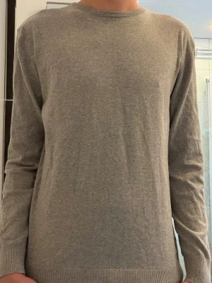 Grå stickad tröja från Indicode - En enkel och stilren grå stickad tröja från Indicode i storlek medium. Tröjan har rund halsringning, långa ärmar och ribbade muddar vid ärmslut och nederkant. Perfekt för lager på lager under höst och vinter.