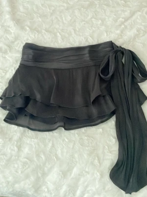 Jättefin Svart kjol - Helt ny, aldrig använd pga fel storlek!! Supersnygg svart kortkjol från Gina 14+ med bred knytband i midjan och dubbla volanger. Kjolen har en flowy känsla och är gjord i ett jättefint glansigt material. Det går att knyta bandet på olika sätt beroende på hur långt ner man vill att det ska hänga ( bild 1 och 3). Baksidan är stretchig i midjan. (Nypris : 329kr)