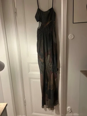 Långklänning NAKD - Säljer en långklänning med tunna axelband och abstrakt mönster i mörka toner som grön, brun och vinröd. Klänningen har flera rynkade sömmar över midjan och är tillverkad i ett tunt, flowy material som ger ett snyggt fall. Använt 1 gång