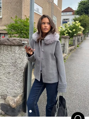 Grå kappa med fuskpäls-krage - Snygg grå kappa med bred, fluffig fuskpäls-krage i ljusgrått. Jackan har knappar framtill, raka ärmar med uppvikta muddar och två fickor. Perfekt för dig som vill ha en stilren och trendig look under kallare dagar. priset går att diskutera, använd 3 gånger då jag köpte en i svart istället 