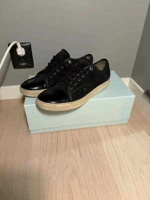 Svarta sneakers från Lanvin med lackdetalj - Säljer ett par svarta Lanvin sneakers med snygg lackad tå och ovandel i mocka. Skorna har svarta snören, metallöljetter och vit sula. Klassisk low top-modell med exklusiv känsla och cool kontrast mellan material. Kommer med originalkartong.