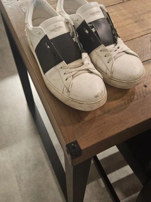 Valentino Open Garavani sneakers (äkthetsbevis) - Säljer ett par Valentino Open Garavani sneakers i vitt och svart skinn med ikoniska nitar runt hälen. Skorna har snörning och platt sula, med en chunky och modern siluett. Medföljer originalkartong, dustbags och extra skosnören. Skorna är i bra skick men lite slitage vid tån på höger sko. Skorna är smutsiga men går att tvätta bort enkelt med ljummet vatten. Pris kan diskuteras! Hör av er om intresserad!