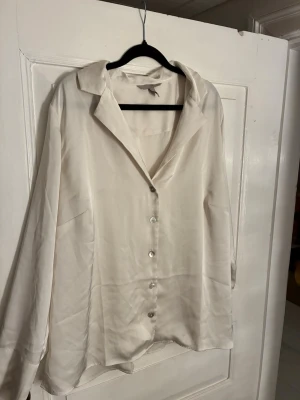 Vit satinliknande skjorta från H&M - Säljer en vit skjorta från H&M i storlek XL. Skjortan har en lyxig satinliknande finish, klassisk krage och knappar framtill. Långärmad modell med breda manschetter och avslappnad passform. Perfekt för dig som gillar stilrena och enkla plagg.