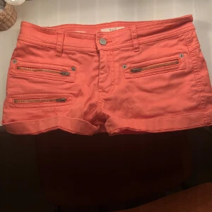 Korallrosa shorts med dragkedjor - Säljer ett par vintage korallrosa shorts med fyra snygga dragkedjor framtill och två baktill. De har uppvikta benslut, bälteshällor och en klassisk knappstängning. Perfekta för sommaren och riktigt sköna att bära till allt. Materialet känns som bomull.