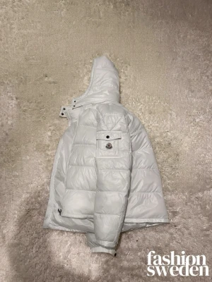 Vit pufferjacka från Moncler - Säljer en riktigt snygg vit pufferjacka från Moncler med huva och dragkedja. Jackan har quiltad design, fickor med dragkedja och en ikonisk Moncler-logga på bröstet. Perfekt för kalla dagar och ger en clean look. Passar dig som vill ha både stil och värme.
