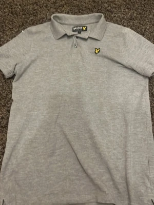 Grå piké från Lyle & Scott - Säljer en riktigt snygg o stilren grå pikétröja från Lyle & Scott. Tröjan har korta ärmar och den ikoniska gula loggan på bröstet. Storlek xs/s. 