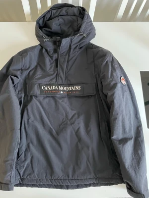 Svart skidjacka från Alcott - Svart skidjacka med huva från Alcott, fodrad med mjuk fleece på insidan. Stor ficka med dragkedja och tryckknappar framtill, samt broderad logga 'Canada Mountains'. Justerbar huva med snörning och diskret logotyp på ärmen. Perfekt för kyliga dagar. Köpt i Italien 🇮🇹 Helt ny utan prislapp.