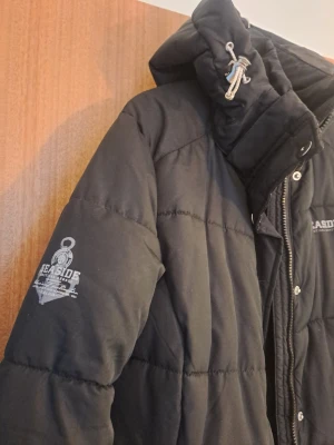 Svart pufferjacka från Seaside - Säljer en svart pufferjacka från Seaside med huva och dragsko. Jackan har quiltad design, dragkedja och knappar framtill samt logga på bröstet och ärmen. Perfekt för kalla dagar och riktigt skön att bära.