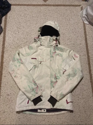 Vit mönstrad vindjacka från Salomon - Säljer en vit vindjacka från Salomon med abstrakt mönster i grönt och lila. Jackan har huva, dragkedja och flera fickor med dragkedjor. Materialet är syntet och jackan har långärmade muddar. Perfekt för dig som gillar att sticka ut med coola detaljer.