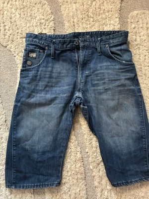Blå jeansshorts från G-Star RAW - Säljer ett par klassiska blå jeansshorts från G-Star RAW med fem fickor, bälteshällor och dragkedja. Shortsen har en avslappnad passform och snygga slitningar för en cool look. Perfekta till sommaren och lätta att matcha med allt. Storlek W32.