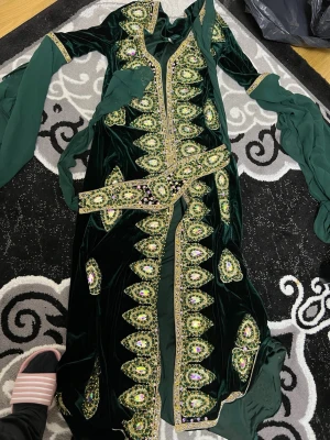 Grön långklänning med paljettdetaljer - Säljer en riktigt maffig långklänning i mörkgrön sammet med glittriga paljettdetaljer och broderier i guld och grönt. Klänningen har långa ärmar och ett matchande bälte i midjan. Perfekt för dig som vill sticka ut och älskar detaljer!