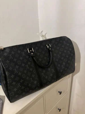 Svart  resväska i skinn - Säljer en svart  resväska i skinn med klassiskt monogrammönster i grått. Väskan har silverfärgade metalldetaljer, dubbla handtag och avtagbar axelrem. Perfekt för dig som vill resa med stil och ha plats för allt du behöver.