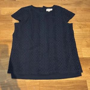 Mörkblå spetsblus från Holly & Whyte - Snygg mörkblå blus från Holly & Whyte med korta ärmar och rund hals. Skriv för bilder hur den sitter på. Blusen har ett vackert spetsmönster över hela plagget och är fodrad undertill. Perfekt att styla med jeans eller kjol för en trendig look. Storlek 146/152