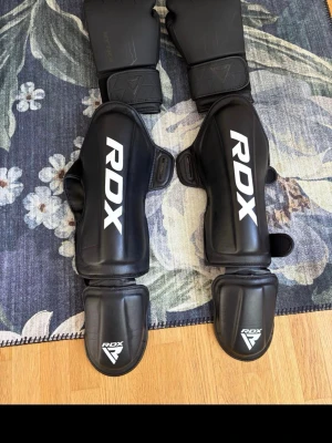 Svarta RDX benskydd och handskar - Säljer ett par svarta benskydd och matchande handskar från RDX, perfekta för kampsport eller träning. Benskydden har bred kardborreknäppning och är tillverkade i slitstarkt syntetmaterial med vit logga. Handskarna är vadderade och har smart öppning för ventilation. Personer mellan 150-170cm kan använda benskydden och boxningshandskarna är 14oz så det är perfekt för sparring och träning. Ganska ny luktar inte svätt. Hela sättet kosta mig 1400kr