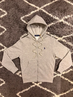 Polo Ralph Lauren Zip hoodie  - Polo Ralph Lauren zip-hoodie Grå hoodie med hel dragkedja, huva med snörning och broderad Polo-logga på bröstet. Mjuk och varm insida. Klassisk och bekväm modell för vardagsbruk. Jag väger 70KG och är 180CM och den är lite liten på mig. . 