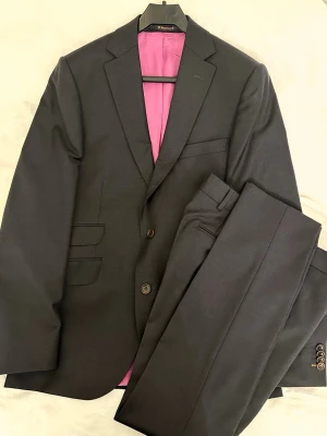 Morris mörkgrå ullkostym strl 46 - Säljer en stilren mörkgrå kostym från Morris i 100% ull, storlek 46. Kavajen har klassisk passform, två knappar och snyggt rosa foder med randiga detaljer på insidan. Byxorna har enkel design med bälteshällor och knappstängning. Väldigt bra skick!