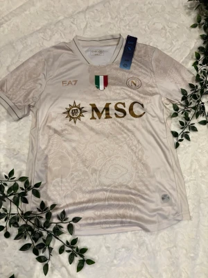 Napoli EA7 fotbollströja vit/guld S - Snygg Napoli matchtröja från EA7 i vitt med gulddetaljer och diskret mönster. Tröjan har klubbmärke, italienska flaggan och MSC-logga framtill. Tillverkad i lätt polyester, perfekt för fotboll eller som streetwear.
