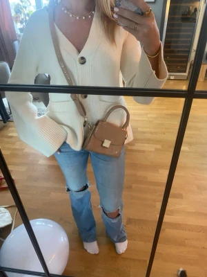 Liten beige väska från Glitter  - Liten stilren väska från Glitter med gulddetalj. Denna passar perfekt till varje tillfälle och adderar en stilren touch till varje outfit. 