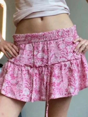 Rosa blommig volangkjol från Zara - Supersöt kort kjol i rosa och vitt med blommigt mönster och volanger. Kjolen har bred resår i midjan och snörning framtill för extra detalj. Lätt och luftig, perfekt för varma dagar. Passar dig som gillar en feminin och avslappnad stil.