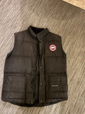 Svart dunväst från Canada Goose - Säljer en svart dunväst från Canada Goose med hög krage, stora framfickor och klassisk logga på bröstet. Västen har både dragkedja och tryckknappar framtill. Perfekt för lager-på-lager och riktigt snygg till höst- och vinterlooken.