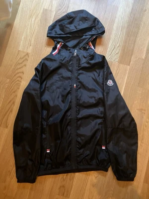 Svart vindjacka från Moncler - Snygg svart vindjacka från Moncler med huva och dragkedja framtill. Jackan har Moncler-logga på ärmen och coola detaljer med röd, vit och blå rand vid huvan. Perfekt för dig som vill ha en stilren och sportig look. Pris går att diskutera vid snabb affär