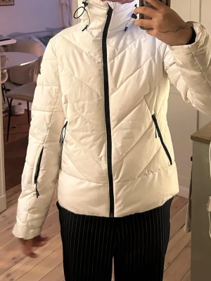 Vit skidjacka från McKinley, strl 38 - Säljer en snygg vit pufferjacka från McKinley i storlek 38. Jackan har huva, svarta dragkedjor och snörning vid kragen. Quiltad design och praktiska fickor med dragkedja. Perfekt för kalla dagar och riktigt skön att bära.