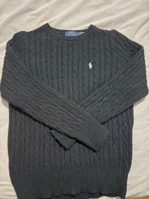 Svart kabelstickad tröja Polo Ralph Lauren - Kabelstickad svart tröja från Polo Ralph Lauren med klassisk rund hals och den ikoniska vita logon broderad på bröstet. Tröjan har lång ärm och ribbade muddar, perfekt för kyliga dagar och en clean look.