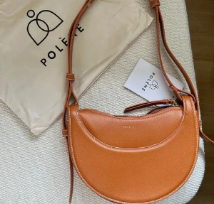 Brun axelväska från Polène i skinn - Säljer en snygg brun axelväska i halvmåneformad design. Väskan är gjord i skinn med justerbar axelrem och har guldfärgad dragkedja. Perfekt för dig som vill ha en stilren och trendig accessoar till vardagslooken. Vet ej om äkta därav priset köpt på plick! 