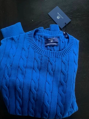 Blå kabelstickad tröja från GANT - Säljer en blå kabelstickad tröja från GANT i premium bomull. Tröjan har rund halsringning, ribbade muddar och klassiskt kabelmönster. Perfekt för dig som gillar en tidlös och fräsch look. Märkets logga broderad på bröstet.
