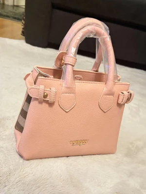 Burberry damväska - Säljer en superfin rosa handväska från Burberry. Den har ett klassiskt rutigt mönster på sidorna och är gjord i skinn. Väskan har två handtag och snygga spänndetaljer på sidorna. Insidan är rymlig med flera fack och en dragkedja för säker förvaring. Perfekt för både vardag och fest