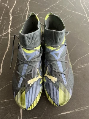 Puma fotbollsskor i grått och gult - Säljer ett par snygga Puma fotbollsskor med högt skaft och snörning. Skorna har en mix av grått, ljusblått och gult med coola grafiska detaljer och Puma-logga på sidan. Ovandelen är i syntetmaterial och sulan har fasta dobbar för bra grepp på planen.