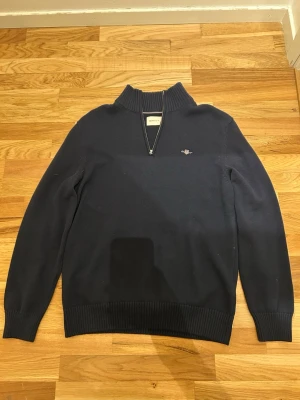 Marinblå stickad tröja från GANT - Säljer en marinblå stickad tröja från GANT i storlek L. Tröjan har ribbad hög krage och half zip-dragkedja framtill. Klassisk GANT-logga broderad på bröstet och långa ärmar. Perfekt till höst och vinter för en clean och stilren look.