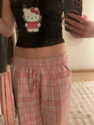 Svart Hello Kitty linnetopp XS - Supersöt svart croppad linnetopp med Hello Kitty-tryck framtill och tunna axelband. Toppen har en liten vit rosett vid urringningen och är perfekt för dig som gillar gulliga och lekfulla plagg. Materialet känns mjukt och stretchigt.