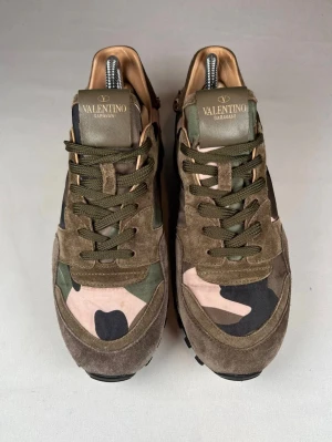 Valentino Rockrunner sneakers camouflage - Snygga Valentino Rockrunner sneakers med camouflage-mönster i grönt, beige, rosa och svart. Ovandel i mocka och textil, med coola nitar runt sulan och grov svart yttersula. Klassisk snörning och rund tå. Perfekt för dig som vill sticka ut med exklusiva sneakers.