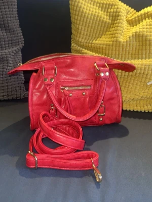 Röd handväska med gulddetaljer - Snygg röd handväska med guldfärgade nitar och spännen. Väskan har dubbla handtag samt en tillhörande axelrem som kan fästas på sidorna. Den har en liten ficka inuti med dragkedja. Köpt i Italien och har aldrig använts. Säljer pga att det inte är min stil.❤️