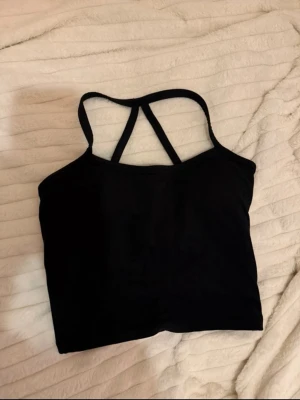 Svart croppad sport topp med smala band - En svart croppad linnetopp med smala axelband som korsas i ryggen. Den är vadderad och i tjockt material.  Strl S, aldrig använd 