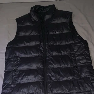 Svart dunväst från Hugo boss - Svart dunväst från Hugo boss med glansig finish och quiltad design. Västen har hög krage, två fickor med knappstängning och en dragkedja framtill. Stort Hugo-tryck i svart på ryggen ger en cool look. Perfekt att slänga över en hoodie för extra värme.