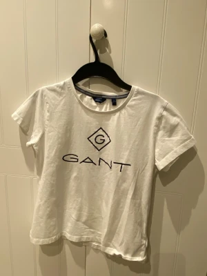 Vit GANT t-shirt med logotryck - Vit t-shirt från GANT i storlek M med klassiskt svart logotryck på bröstet. Rund hals och korta ärmar, tillverkad i mjuk bomull som känns skön mot huden. Perfekt för dig som gillar stilrena och enkla plagg med tydlig branding.