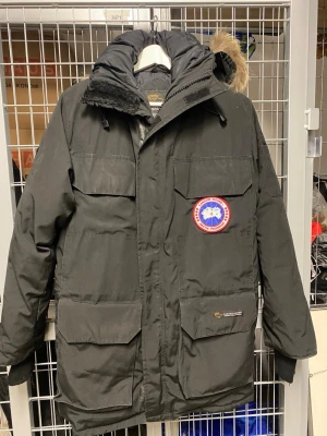 Svart Canada Goose Expedition dunjacka - Säljer en svart Canada Goose Expedition dunjacka med stor huva och äkta pälsdetalj. Jackan har flera stora fickor, patch på bröstet och Expedition-märke på ryggen. Perfekt för kalla vinterdagar och riktigt varm. Klassisk och tidlös modell.