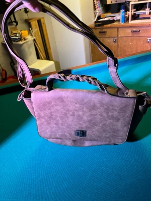 Brun axelväska med flätat handtag - Säljer en brun axelväska i skinnimitation med flätat handtag och justerbar axelrem. Väskan har ett lock med metallspänne och ett rymligt innerfack med färgglatt paisleymönstrat foder och dragkedja. Perfekt för dig som vill ha en snygg och praktisk väska.