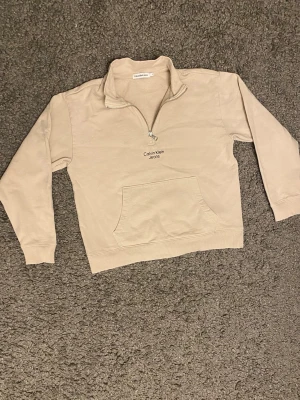 Beige half zip tröja Calvin Klein Jeans - Snygg beige långärmad tröja från Calvin Klein Jeans med half zip och broderad logga på bröstet. Tröjan har en känguruficka framtill och är gjord i mjuk bomull. Perfekt för chill dagar och enkel att matcha med jeans eller joggers.